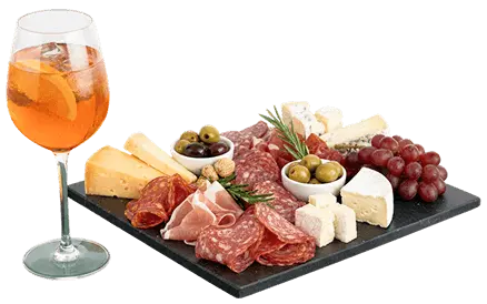 un calice di Spritz poggiato vicino a un tagliere in ardesia con sopra vari salumi, formaggi, olive e uva, disposti in modo invitante